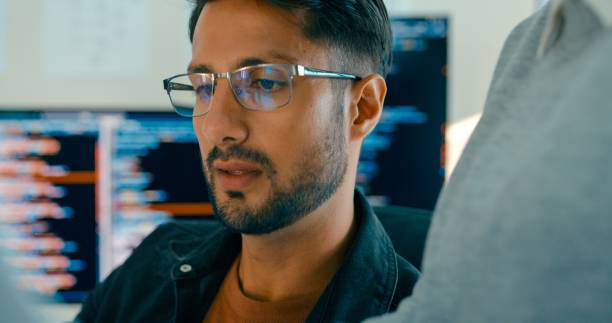 Мужчина в очках работает над программированием за компьютером с несколькими экранами на заднем плане.
